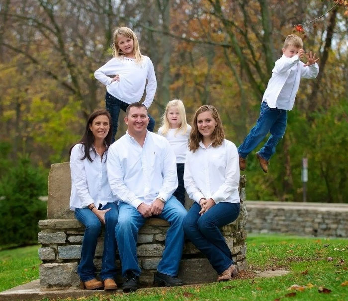 Family posing outdoors in white shirts, with a child humorously jumping off a stone bench in a park.