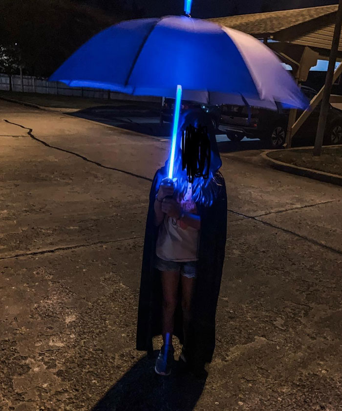 Child wearing a cape holding a glowing blue light sword and umbrella at night, perfect for unique gifts they didn’t see coming.