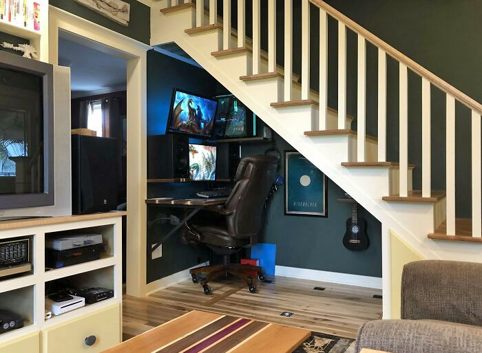 Cozy home office design under staircase with dual monitors, leather chair, guitar, and dark green walls in a small space.