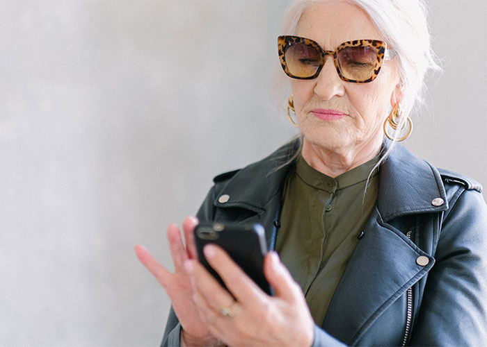 Older woman wearing sunglasses and a leather jacket, focused on her smartphone screen indoors, reacting to paternity test results.
