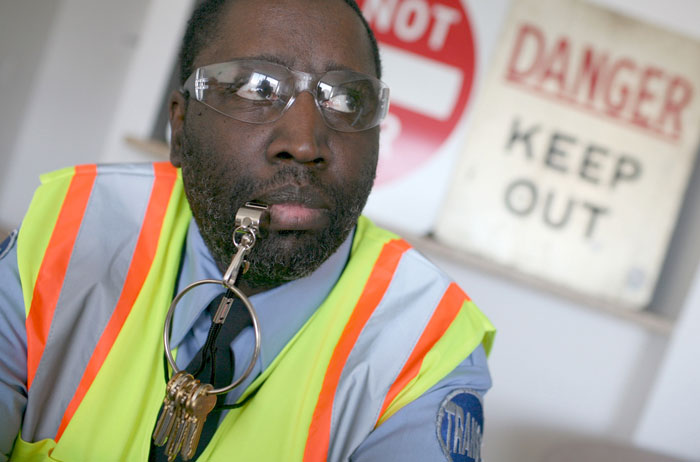 Security officer with a whistle and keys, wearing a safety vest, standing in front of warning signs.
