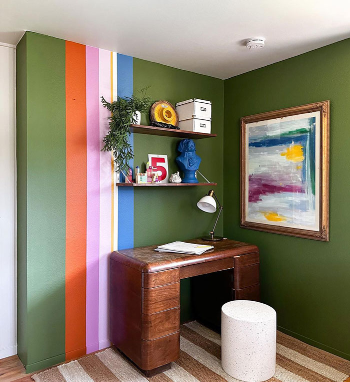 Colorful strips wall painting design on green walls in a cozy workspace with wooden desk and decorative shelves.