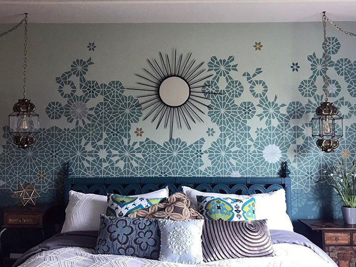 Stencil wall painting design with patterned floral shapes in blue and gold above a bed in a living room setting