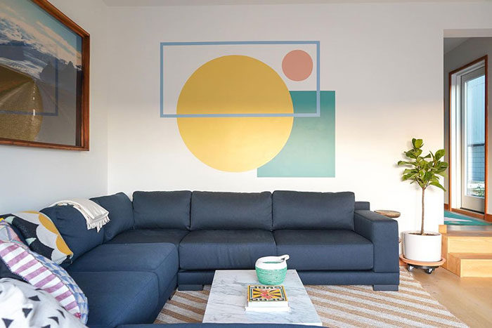 Geometric wall painting design with large shapes in yellow, green, and pink above a dark blue sectional sofa in a living room.