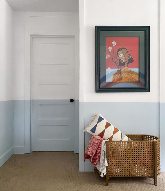 Two-toned wall painting design in soft blue and white with a framed abstract portrait and woven basket decor.