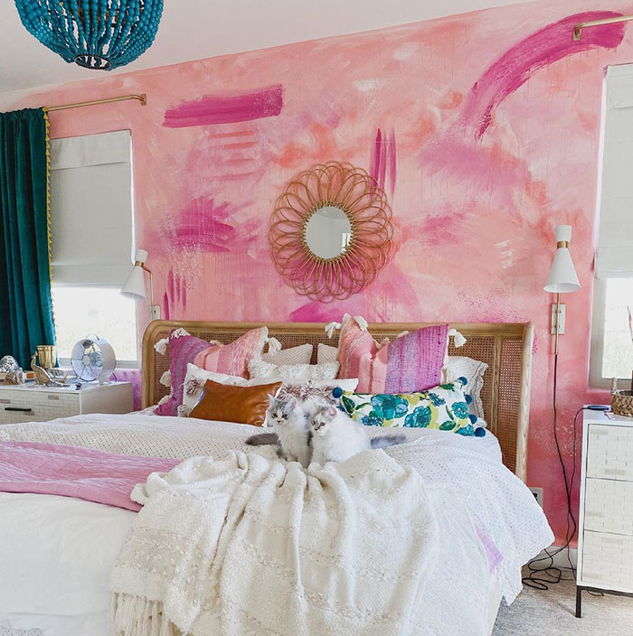 Watercolor wall painting design in bedroom with pink hues, colorful pillows, cozy bedding, and two cats on the bed.