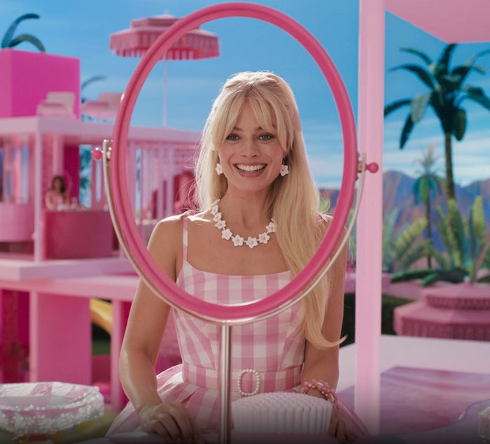 Blonde woman in a pink checkered dress smiling, sitting in a brightly colored room with a large ring-shaped mirror.