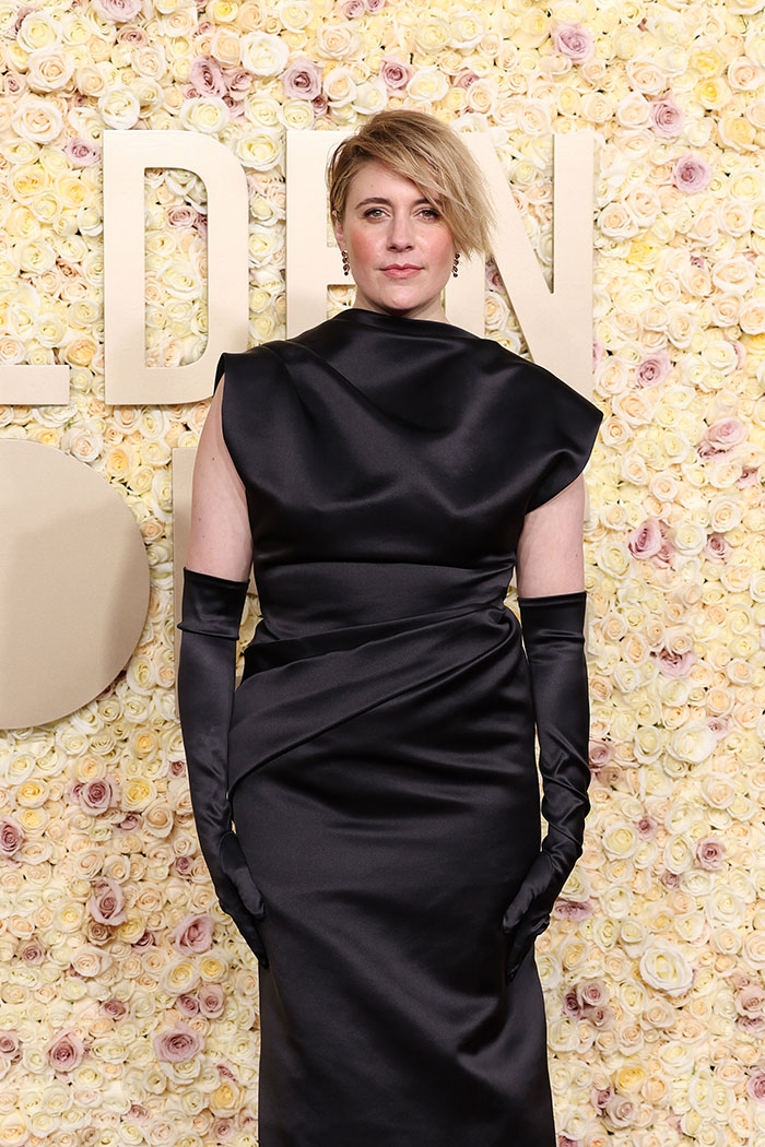 Greta Gerwig in a black dress, standing in front of a floral backdrop at an event.