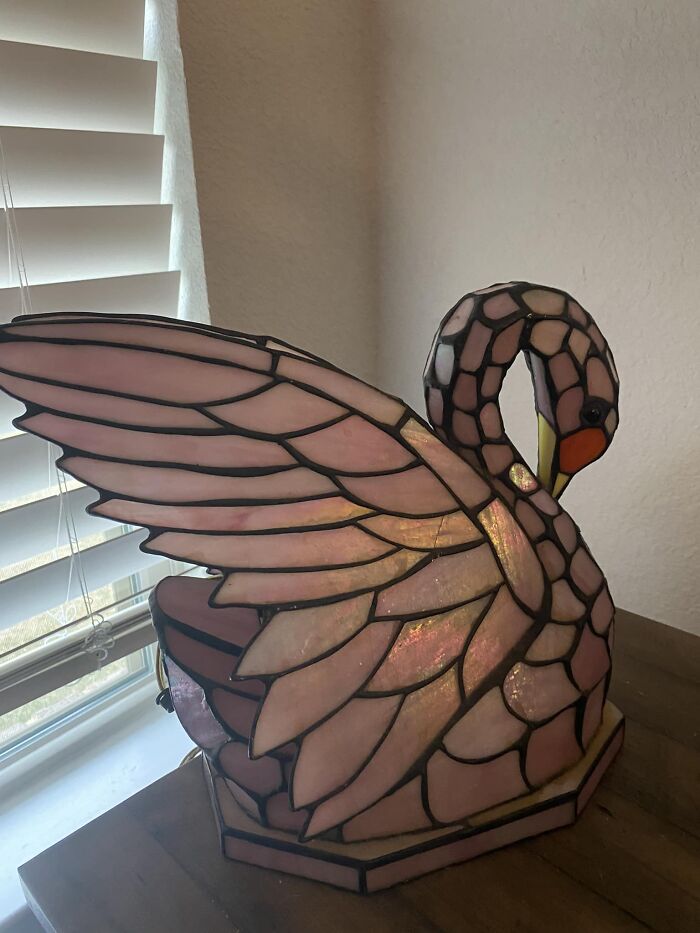 Stained glass swan lamp found at thrift store with intricate pink and orange design, resting on wooden surface near window blinds.