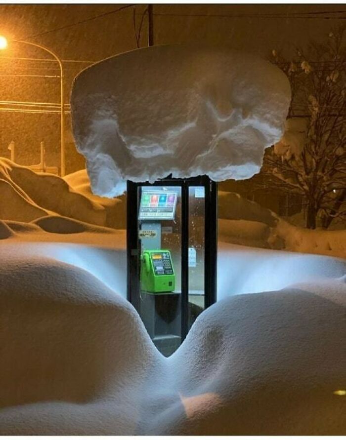 A phone booth covered in heavy snow, illuminated at night, creating a funny and surreal scene.