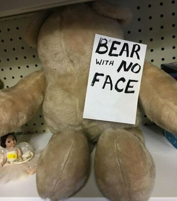 Faceless teddy bear on shelf with a playful sign humorously reading "BEAR WITH NO FACE".