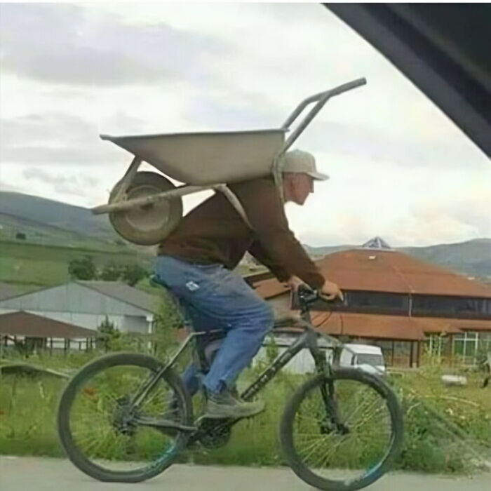 Man riding a bicycle with a wheelbarrow strapped to his back, creating a humorous scene without context.