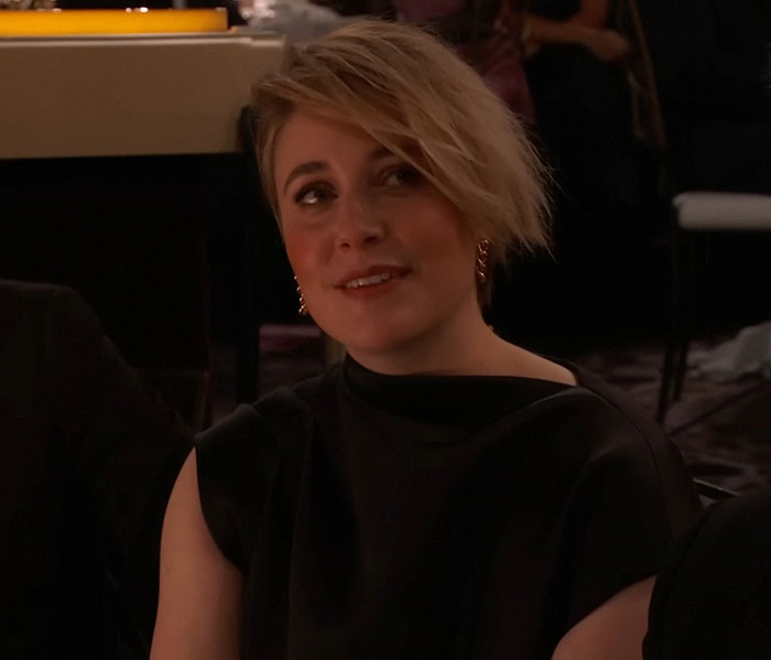 Greta Gerwig in a dimly lit setting, wearing a black outfit, responding to a statement with a thoughtful expression.