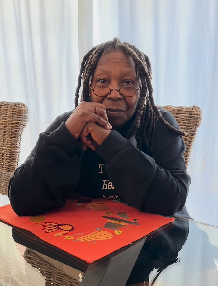 Person sitting at a glass table with a thoughtful expression, wearing glasses and a dark shirt, near wicker chairs.