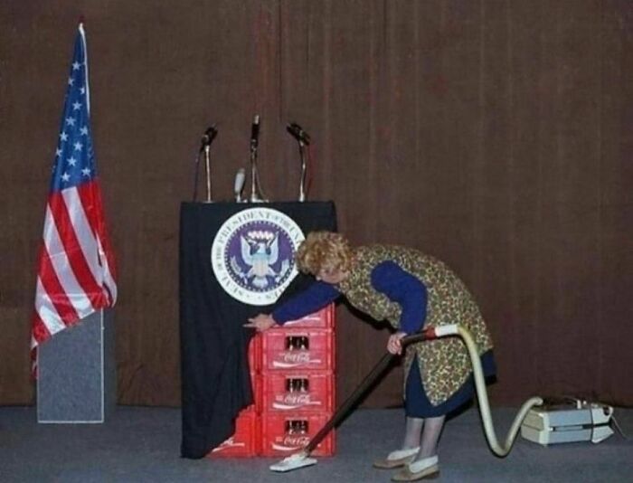 Woman vacuuming near podium with Coca-Cola crates, humorous context from FB page.