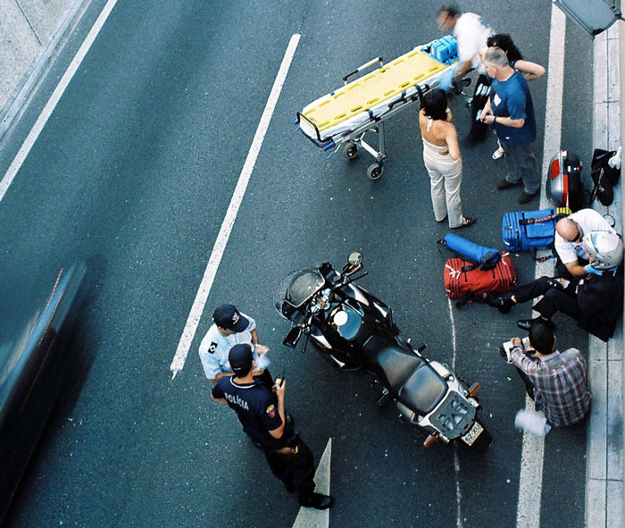 Emergency responders and strangers assisting at a roadside incident involving a motorcycle on a busy street.