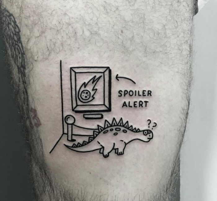 Cartoon dinosaur tattoo with meteor artwork labeled "Spoiler Alert" on pale skin.