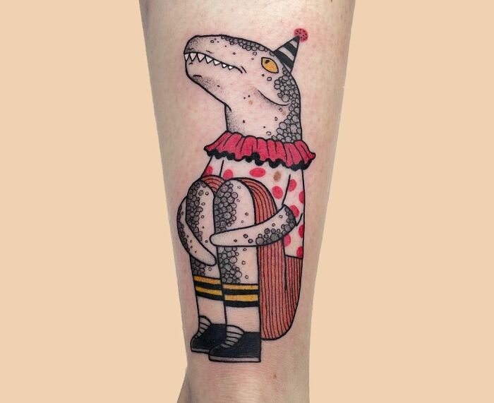 Creative dinosaur tattoo with party hat and polka-dot outfit on forearm.