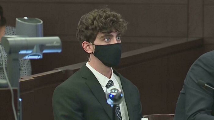 Young man in a courtroom wearing a mask and suit, appearing anxious in a tense and disturbing setting.