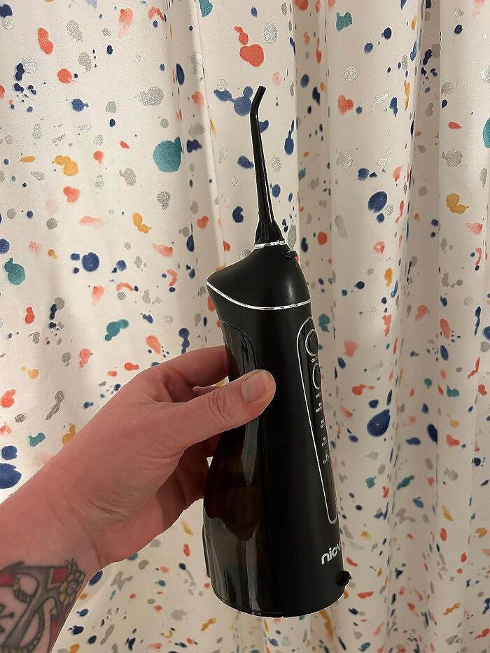 Hand holding a black water flosser in front of a colorful speckled shower curtain, a useful life hack tool.