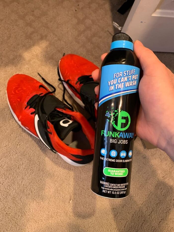 Hand holding odor eliminator spray next to bright orange and black athletic shoes on carpeted floor.