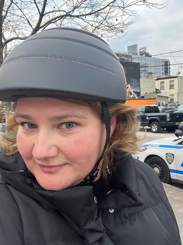 Woman wearing a black helmet and coat outdoors with city buildings and a police car in the background, illustrating life hacks.