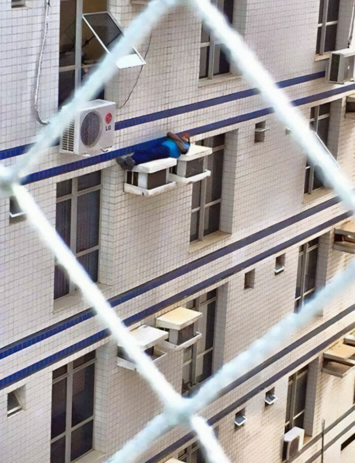 A person lying on a narrow ledge outside a building window, creating a funny and risky scene.