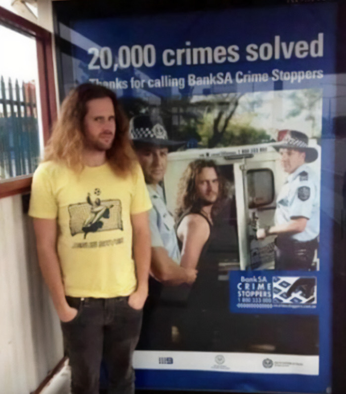 Person in yellow shirt stands by police poster, featuring lookalike in surprising context; image shared on FB page.