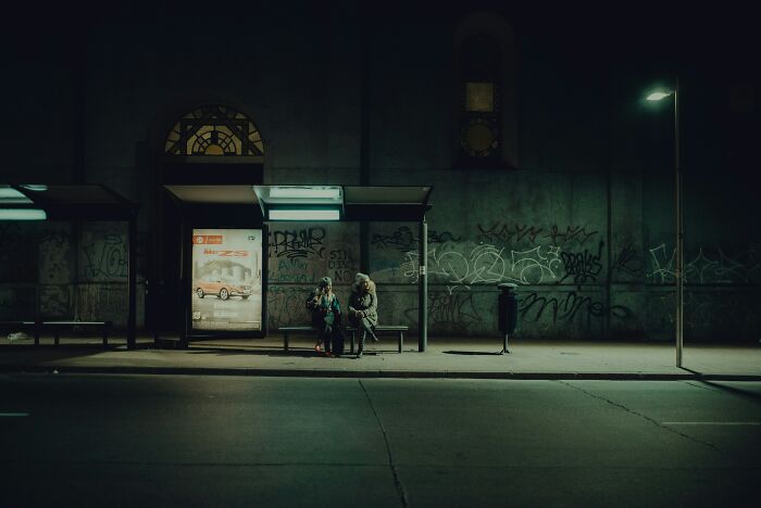 Two people sitting at a dimly lit bus stop at night, evoking stories about incredibly memorable strangers.