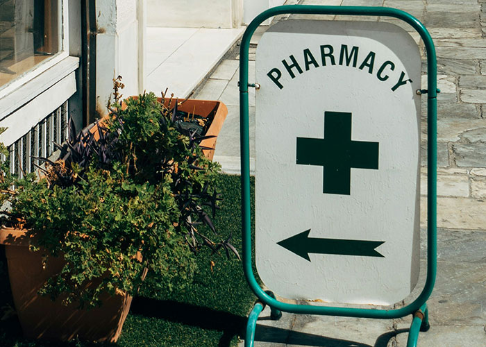 Pharmacy sign with humorous direction arrow, placed near green plants on a sidewalk.
