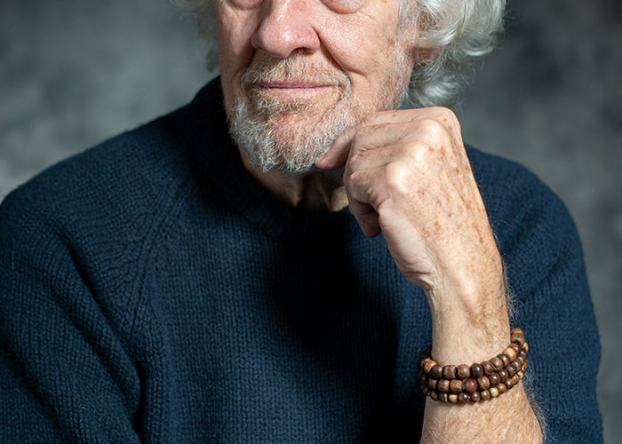 A thoughtful elderly man with white hair, wearing a navy sweater, rests his chin on his hand, humor bracelet on wrist.