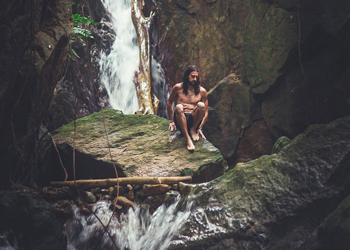 A person sitting on a rock near a waterfall, surrounded by nature, creating an amusing scene despite the serene setting.