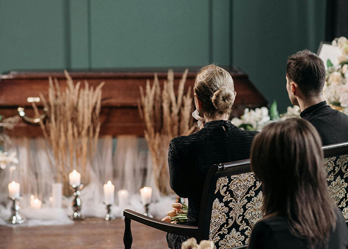 People sitting near a decorated casket at a funeral setting, evoking a humorous context despite the somber event.
