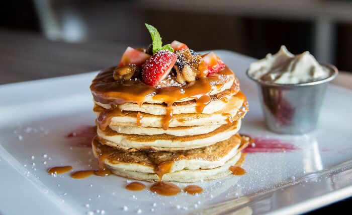 Stack of pancakes topped with caramel, strawberries, and a mint leaf, with whipped cream on the side.