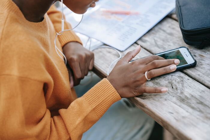Person in an orange sweater scrolling on a smartphone at a wooden table, wearing earphones.