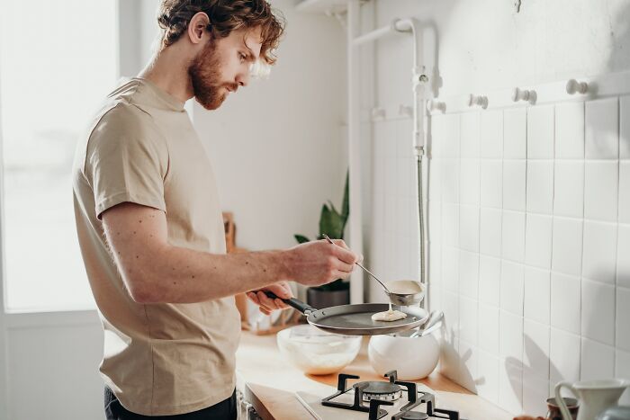 Man in kitchen attempting to cook pancakes, showcasing his struggle with basic cooking skills.
