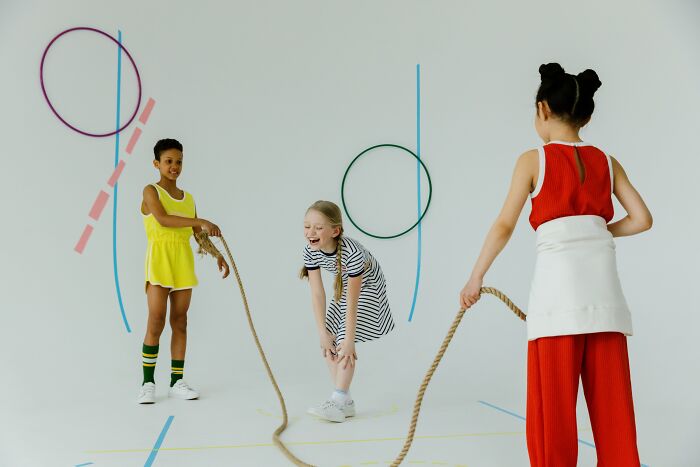 Children playing jump rope, smiling and having fun indoors, with basic gym equipment on walls.