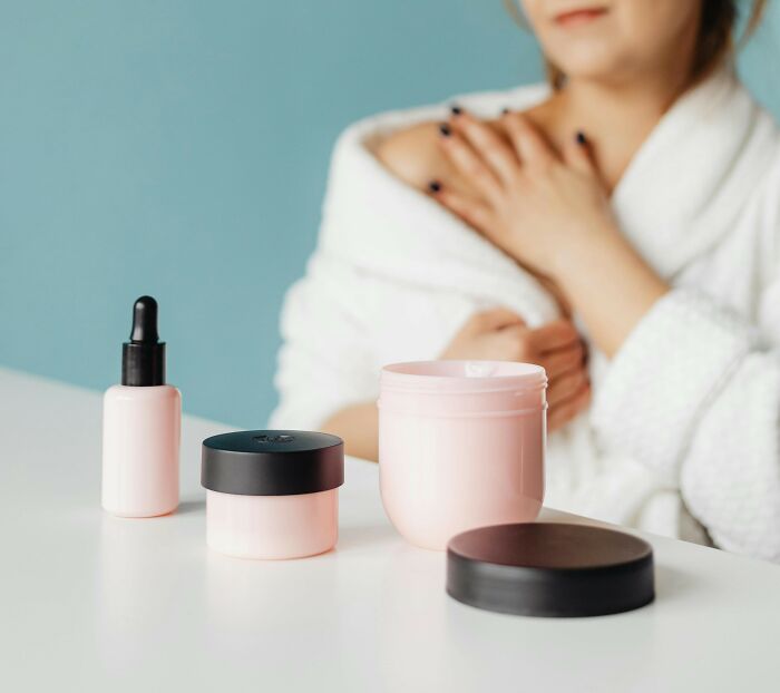 Woman in robe applying lotion, with skincare products on the table.