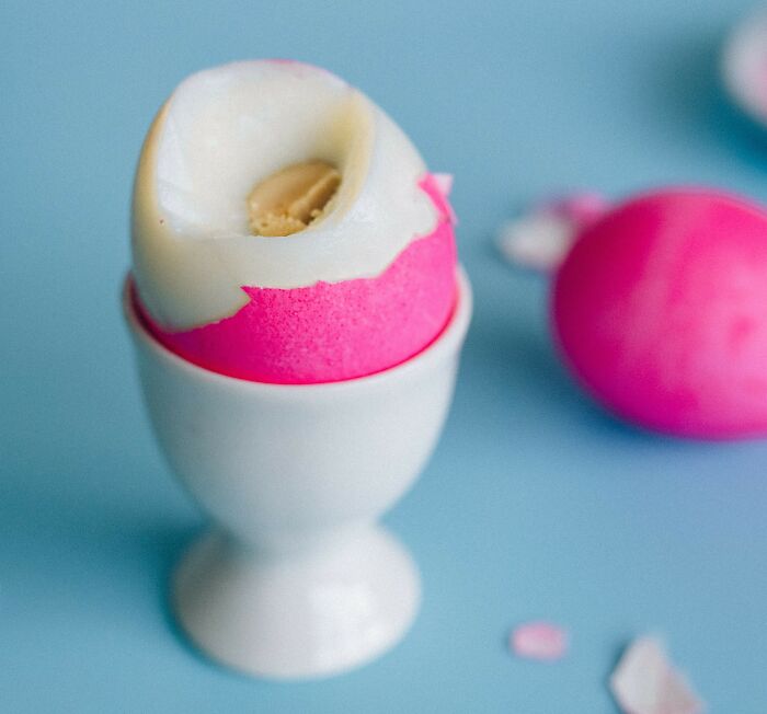 A poorly peeled soft-boiled egg in an egg cup with pink dye on a blue background.