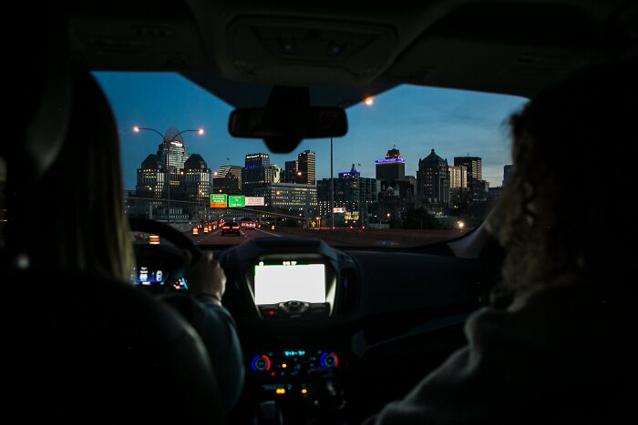 Two people driving at dusk with a city skyline visible through the windshield, capturing memorable strangers moments.
