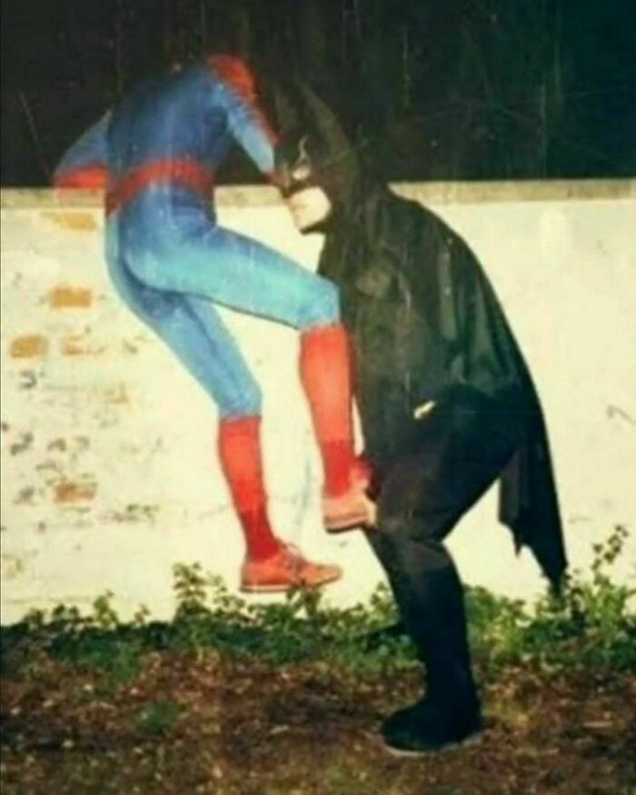 Two people in superhero costumes humorously reenact a scene in front of a wall.