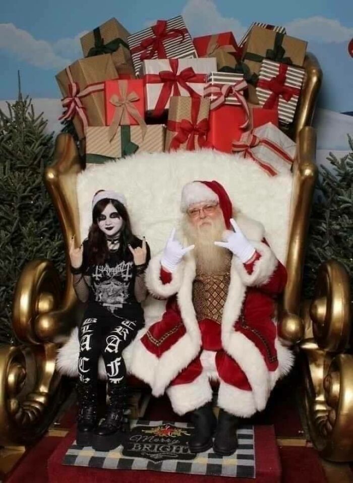 Santa and a person dressed in goth attire make rock hand signs, seated on a chair with gifts, depicting hilarious contrast.