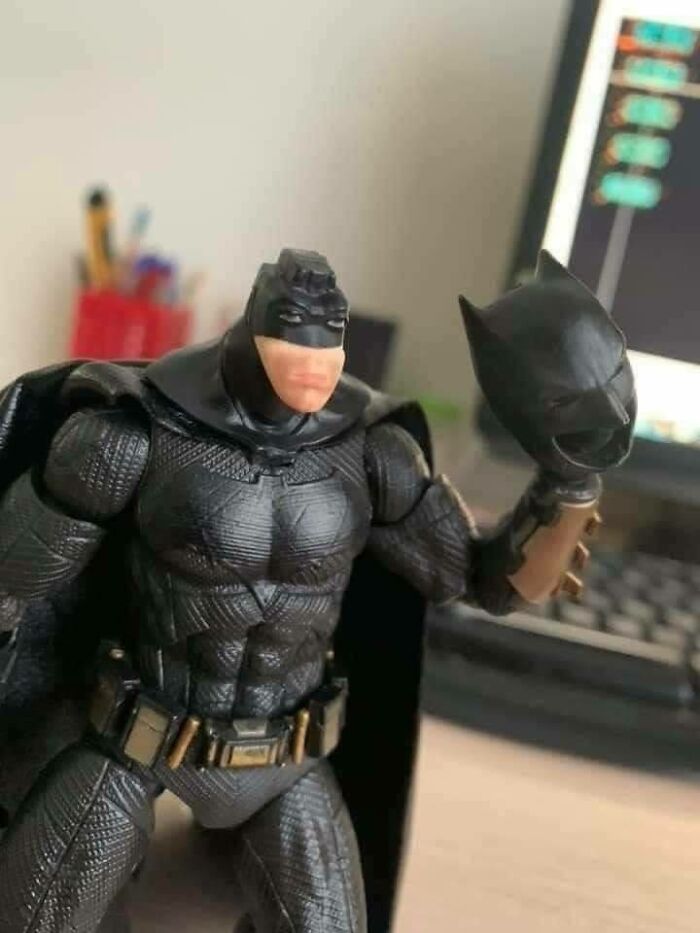 Batman toy with a surprising face holding his mask, creating a humorous scene at a workspace.