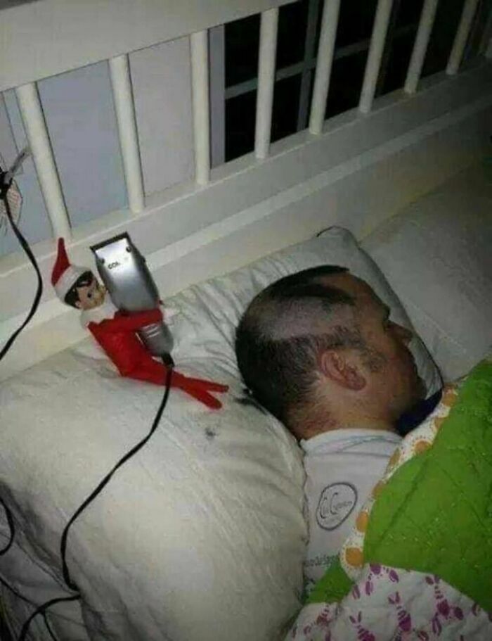 Elf toy holding trimmer beside sleeping man, creating an amusing haircut.