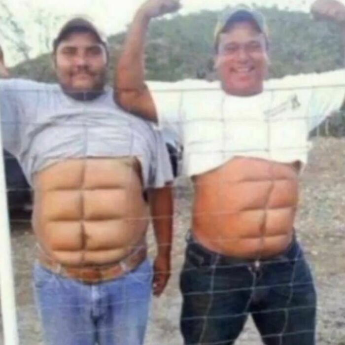 Two men smiling and flexing behind a wire fence, appearing to have defined ab muscles.