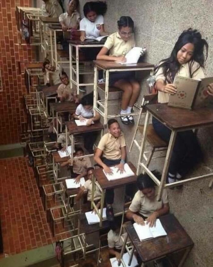 Students hilariously sit at desks arranged vertically on a wall.