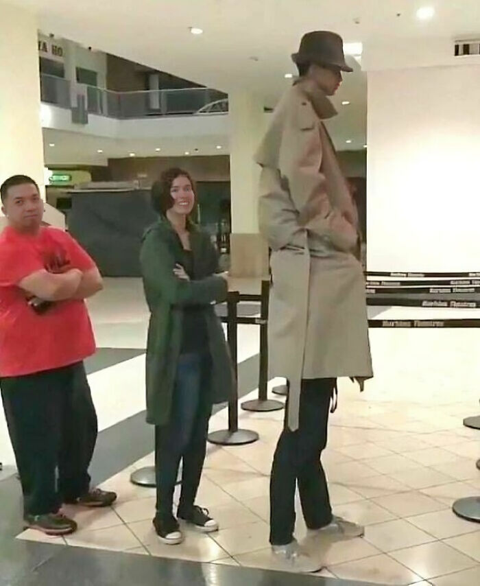 Person in a long trench coat and hat, humorously resembling a classic disguise, stands in line at a public place.