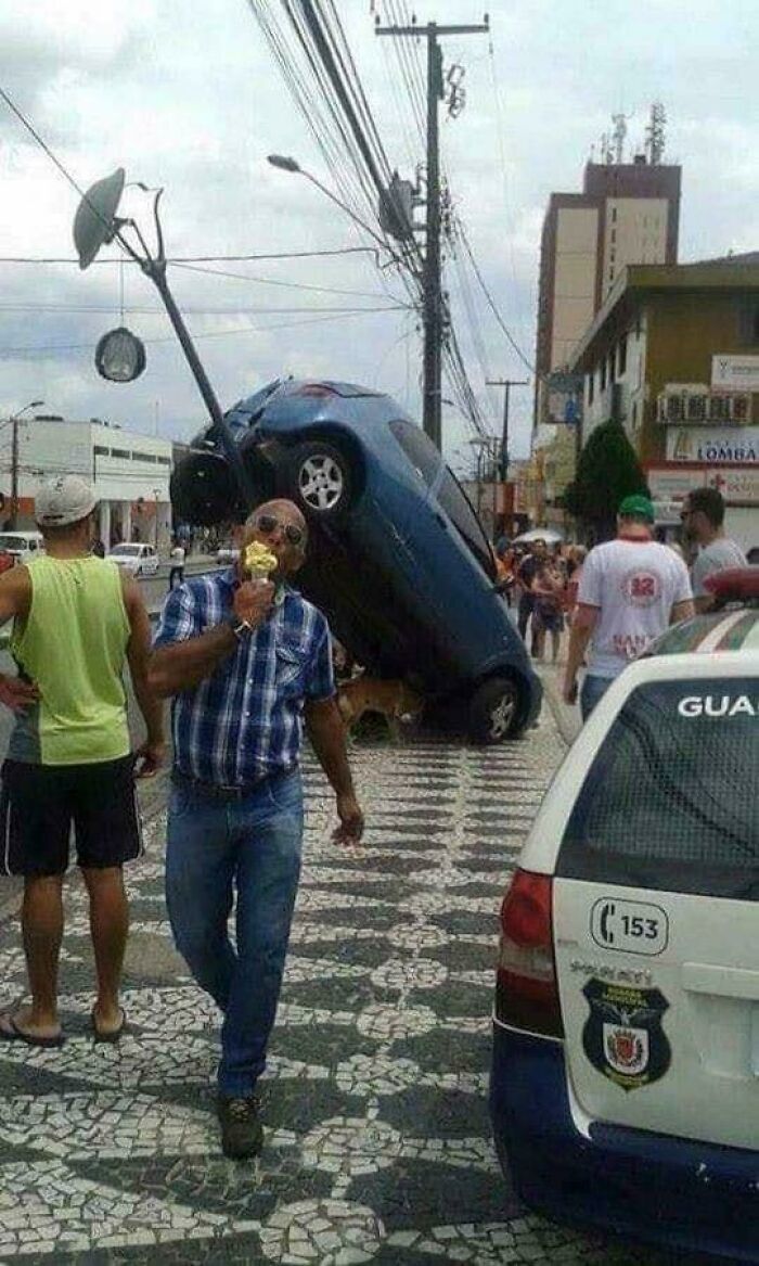 Man eating ice cream with a pole-impaled car in the background, illustrating a funny scene without context.