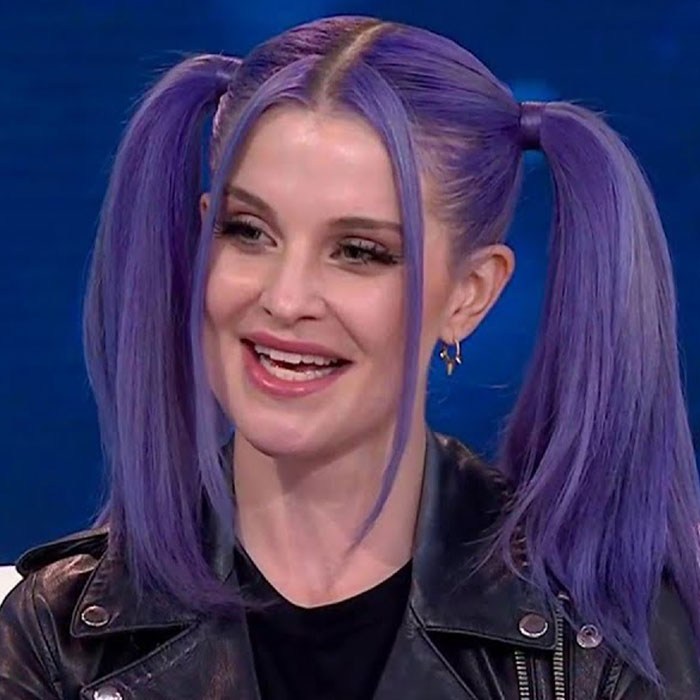 A celebrity with purple hair and leather jacket smiling on a talk show set.