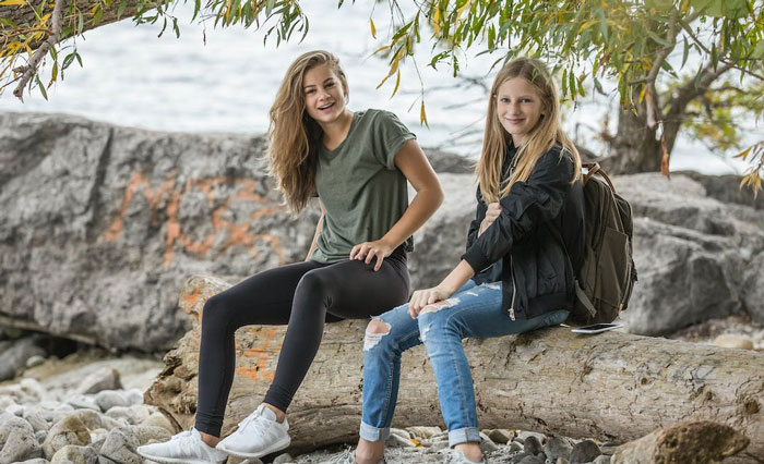 Two girls sitting on a log by the water, enjoying nature and sharing weird, fun facts outdoors.